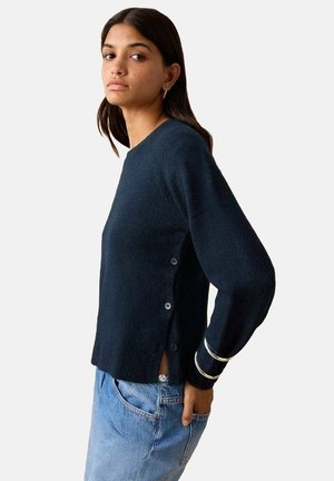 Ženska, oblečena v temno moder pulover z gumbi na strani in svetlo modre jeans hlače, ki pozira z eno roko v žepu na belem ozadju.