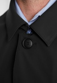 Schwarzer Trenchcoat mit hohem Kragen, ausgestattet mit einem runden schwarzen Knopf und subtilen Stichdetails, über einem hellblauen Hemd geschichtet.