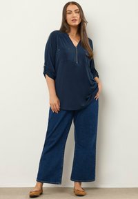Dunkelbluse mit einem Reißverschluss am Ausschnitt, zwei vorderen Taschen und hochgerollten Ärmeln, kombiniert mit weiten, dunklen Jeans und braunen Ballerinas.