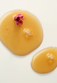 Złote, płynne krople o gładkiej teksturze, z małym różowym kwiatkiem i bąbelkami, na białym tle.