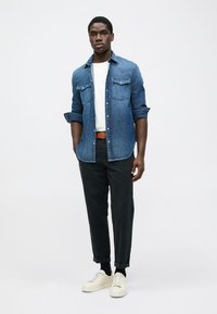 Camicia di jeans di color blu scuro con bottone frontale e tasche sul petto, abbinata a pantaloni neri a gamba affusolata e sneakers color crema. Canottiera bianca semplice visibile.