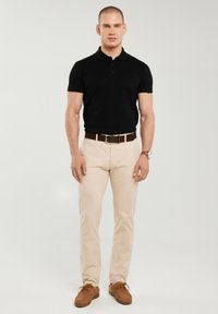 Férfi áll, fekete galléros pólót, bézs nadrágot, barna övet, barna cipőt visel, a bal csuklóján karórával, egyszínű háttér előtt.