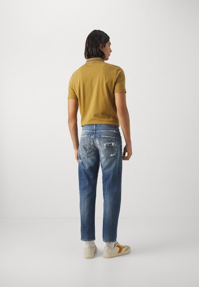 Polo de color verde oliva con mangas cortas, combinado con jeans azules desgastados que presentan detalles rasgados y zapatillas blancas con acentos naranjas.