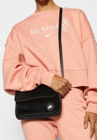 Person som bär en rosa "Be kind" Nike sweatshirt och byxor, håller en svart Nike axelväska med klaff och logopatch.