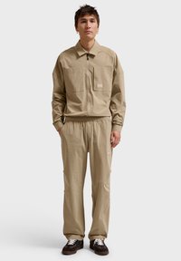 Lys beige, langærmet zip-up top med en lomme, parret med matchende bukser med elastisk talje. Brune og hvide sneakers komplementerer outfittet.