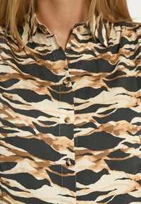 Camicia button-up con stampa animale nei colori beige, marrone e nero. Presenta un colletto, maniche corte e una texture liscia.