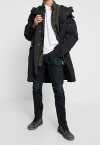 Man i svart parka, grön rutigt skjorta, vit T-shirt, svarta byxor och grå stövlar står mot en enkel ljus bakgrund.