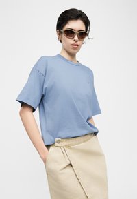 Άτομο με κοντά σκούρα μαλλιά φοράει μπεζ φούστα τύπου wrap, ανοιχτό μπλε μπλουζάκι και οβάλ καφέ γυαλιά ηλίου, στέκεται με τα χέρια στις τσέπες σε λευκό φόντο.