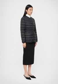 Kvinne iført svart quiltet jakke med knapper over hvit høyhalset genser, svart midiskjørt og svarte spisse slingback hæler, stående sidelengs.