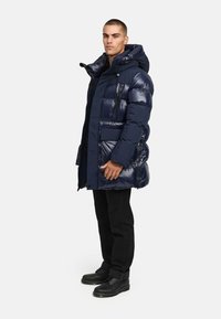 Navy sininen puffatakki huoodilla, jossa on kiiltävä pinta, vaakasuora tikkaus, useita taskuja ja vetoketjusulku. Mustat housut ja saappaat.