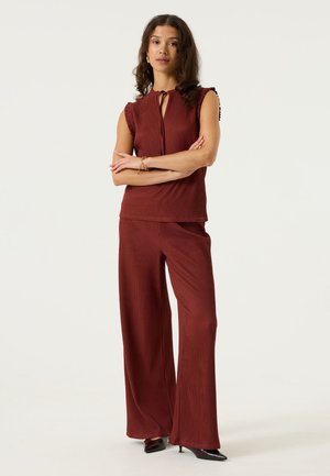 Femme debout, les bras croisés, portant un haut plissé sans manches couleur rouille et un pantalon large assorti, talons noirs, fond neutre.