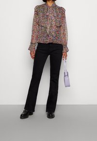 Bloemenachtige doorschijnende blouse met gerimpelde kraag, voorzien van kleine roze en paarse bloemen op een zwarte achtergrond. Gecombineerd met zwarte uitlopende jeans en een lavendelkleurige tas.