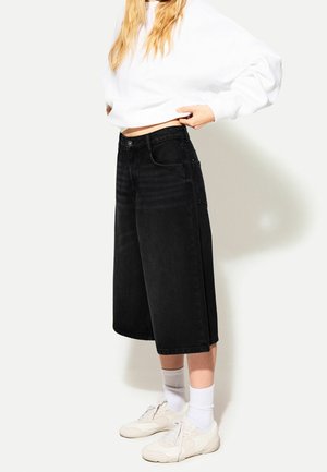 Person trägt ein weißes langärmeliges Oberteil, schwarze, knieumspielende, weit geschnittene Jeansshorts, weiße Knöchelsocken und weiße Sneakers und steht seitlich.