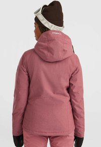 Persoon met een roze ski-jack van O'Neill, bijpassende broek, een bruine gebreide muts, witte skibril en zwarte handschoenen, die met de rug naar ons gekeerd staat.