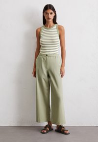 Vrouw in een mouwloos groen-wit gestreept topje, lichtgroene wijde broek en zwarte slide sandalen, staand tegen een witte muur.
