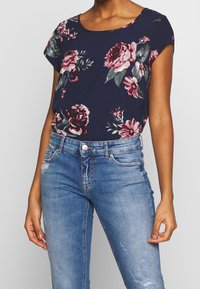 Top floreale navy a maniche corte, con stampe di rose rosse e pink, abbinato a jeans azzurri sbiancati e un dettaglio con bottone argentato.