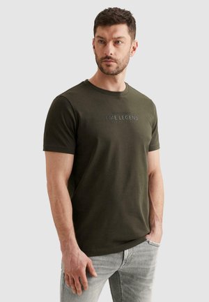 Man draagt een olijfgroene PME Legend T-shirt en lichtgrijze spijkerbroek, staat met één hand in de zak en kijkt iets opzij.