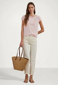 Mulher vestindo uma blusa de manga curta rosa clara, calças de cintura alta off-white, sandálias castanhas com tiras, segurando uma grande cesta de palha.