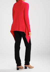 Fuchsiový cardigan s dlouhým rukávem a asymetrickým lemem, s krajkovými detaily na zadní straně. V kombinaci s černými kalhotami a béžovými lodičkami.