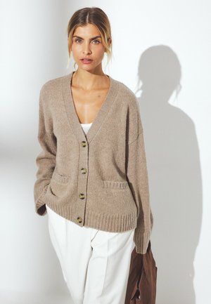 Beige cardigan with a v-neck, button closure, and two front pockets. Ribbed texture and cuffs. Paired with white pants and a brown bag.