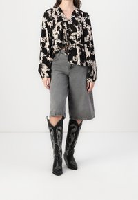 Schwarz-weiß gemusterte Bluse mit langen Ärmeln und einer Schleife in der Taille, kombiniert mit grauen Shorts mit weitem Bein und kniehohen schwarzen Stiefeln mit silberner Verzierung.