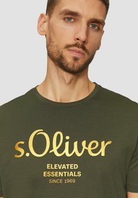 Olivově zelené tričko s ribovanou texturou, na kterém je velké zlaté logo "s.Oliver" a text "NA JEDNÉ ÚROVNI OD ROKU 1969" pod ním. Krátké rukávy.