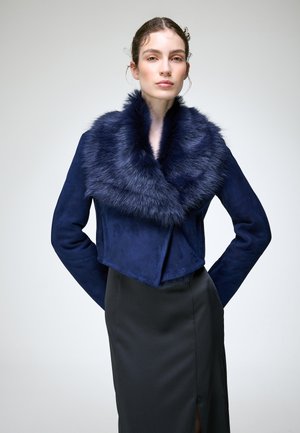 Jeune femme aux cheveux plaqués en arrière portant une veste bleu marine avec col en fourrure et une jupe sombre taille haute, debout devant un fond clair uni.