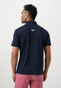 Tumši zils polo krekls ar apkakli un īsām piedurknēm, uz muguras ar baltu logotipu. Gluda tekstūra, piegādājams fit. Kombinēts ar rozā šortiem.