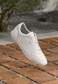 Sneaker en cuir blanc avec une finition texturée, doté d'un bout arrondi, d'une fermeture à lacets et d'un logo discret sur le côté. Semelle en caoutchouc légère.