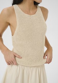 Krämfärgad stickad linne med en texturerad, öppen väv. Har breda axelband och en figurnära bodice, lagd över en flödande beige kjol.