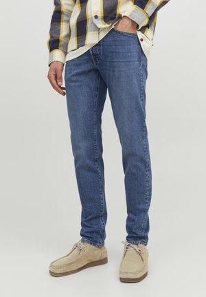 Blaue Denim-Jeans mit schmaler Passform, sichtbaren Nähten und leichter Abnutzung; kombiniert mit beigen Wildlederschuhen und einem karierten Hemd, das über einem weißen Oberteil getragen wird.