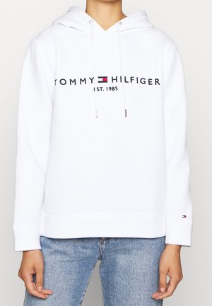 Biała bluza z kapturem z napisem „Tommy Hilfiger Est. 1985” i logo na piersi, noszona przez osobę w niebieskich dżinsach, ręce swobodnie opuszczone wzdłuż ciała.