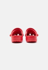 Ζευγάρι κόκκινα καθημερινά λάστιχα τύπου clog με λουράκια για τη φτέρνα, τοποθετημένα σε απλό ανοιχτό γκρι φόντο, που φαίνονται από πίσω.