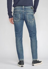 Blauwe spijkerbroek met een fitted ontwerp, subtiele slijtage, achterzakken met stikdetails en versleten accenten. Draag deze met zwarte hoge sneakers.