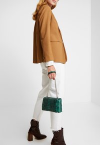 Brun blazer, hvid jeans, mørkebrune hæle støvler. Grøn krokodilleskind prægning håndtaske med kæde rem. Glatte teksturer og struktureret design.