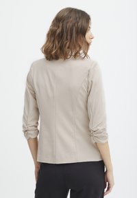 Beige, långärmad blus med rynkade manschetter och en figurnära silhuett. Slätt tyg med en ren, skräddarsydd bakseamsdetalj.