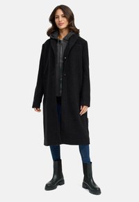Manteau noir en mélange de laine avec un design ajusté, doté d'une couche intérieure grise à capuche. Porté sur un jean sombre et des bottes noires épaisses.