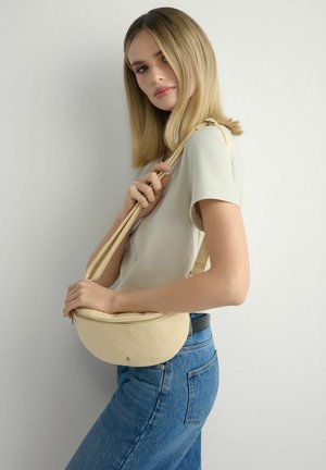 Młoda kobieta o prostych blond włosach, ubrana w beżową bluzkę i niebieskie dżinsy, trzyma kremową pikowaną torebkę na ramię.