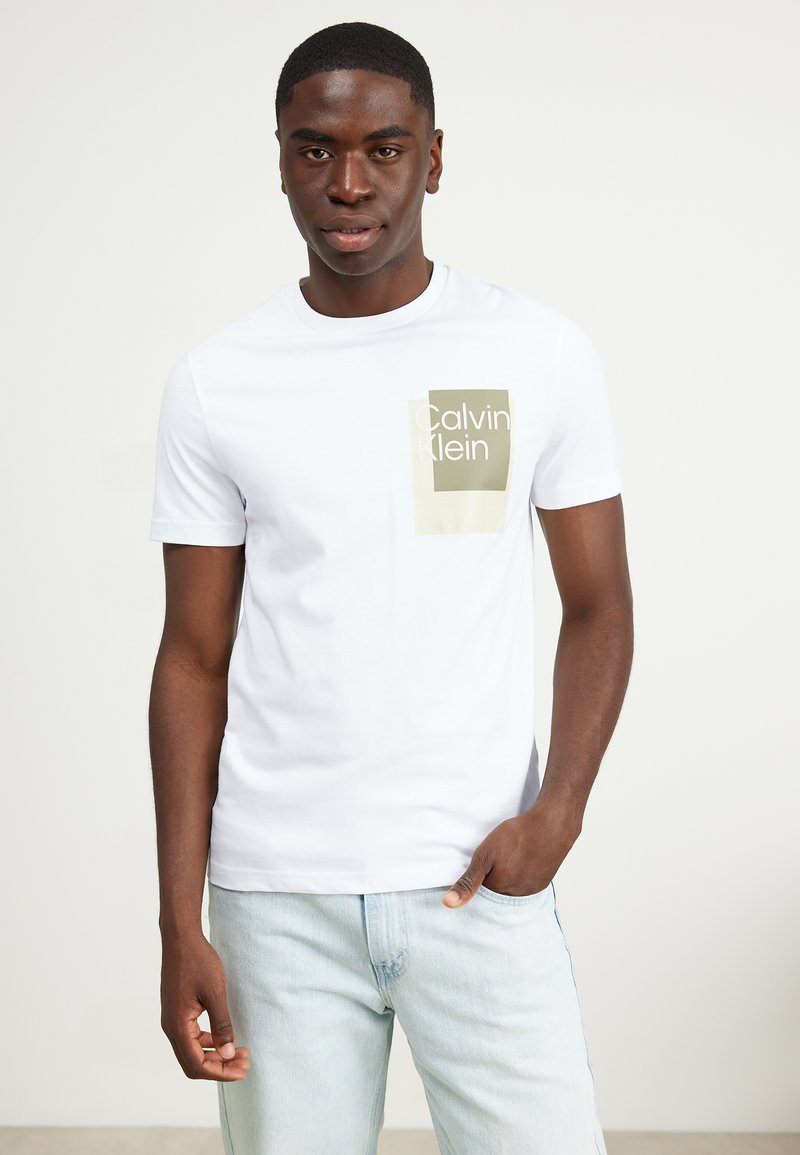 Hvit bomulls t-skjorte med et rektangulært olivengrønt logodesign som leser "Calvin Klein" på venstre brystområde. Korte ermer, rund hals.