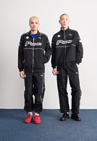 Sorte atletiske jakker og bukser med hvite detaljer og Puma-merkevisning, vist på to individer som står mot en nøytral bakgrunn.