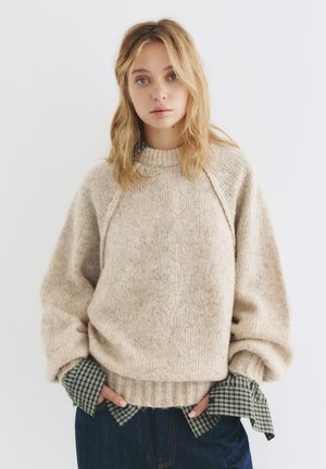 Pull en maille beige avec un ourlet côtelé et des manches raglan. Superposé avec une chemise à carreaux verte aux poignets. Motif en laine texturé visible.