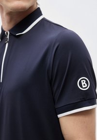 Homme portant un polo bleu marine à manches courtes avec un liseré blanc au niveau du col et des manches, et un logo "B" blanc cerclé sur la manche.