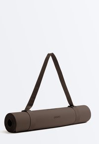 Sötétbarna, feltekerve, vállpánttal ellátott jóga matrac, vízszintesen egy egyszerű világos háttéren fekve.