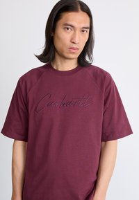 Burgunderrøde t-skjorte med kort erm som har en ribbet rund hals og brodert "Carhartt"-logo i en litt mørkere nyanse. Glatt stofftekstur.
