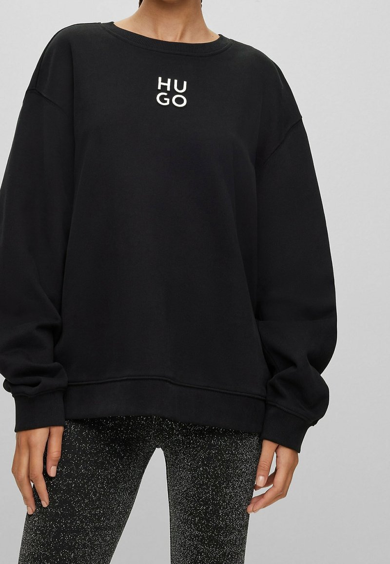 Personne portant un sweat-shirt noir oversize avec le logo "HUGO" sur la poitrine et des leggings noirs tachetés sur un fond gris uni.