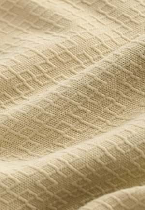 Tissu texturé beige présentant un motif géométrique de diamants en relief et des variations subtiles dans le tissage, avec une apparence douce et lisse.
