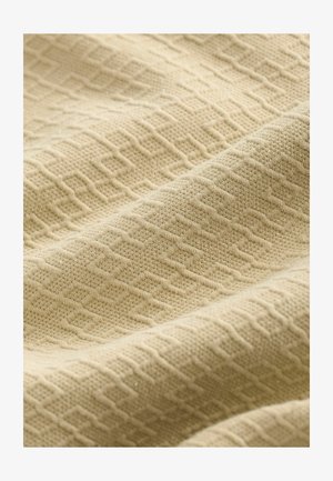 Beige tekstuuriga kangas, millel on geomeetriline mustrid üles tõstetud teemantidega ja peenete kudumisvariatsioonidega, millel on pehme ja sile välimus.