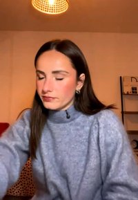 Femme aux longs cheveux sombres et boucles d’oreilles en anneau, portant un pull gris, assise à l’intérieur sous une lumière de plafond ronde, regardant vers le bas les yeux fermés.