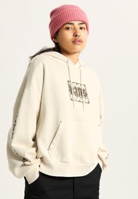 Creme hoodie met een voorzak en grafische print. Beschikt over mouwen met boorden en een geribbelde zoom. Draag het met een roze geribbelde beanie.