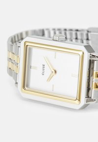 Silberne und goldene rechteckige Uhr mit einem Metallband, ausgestattet mit einem gebürsteten Silberzifferblatt, einfachen Stundenmarkierungen und Logo.